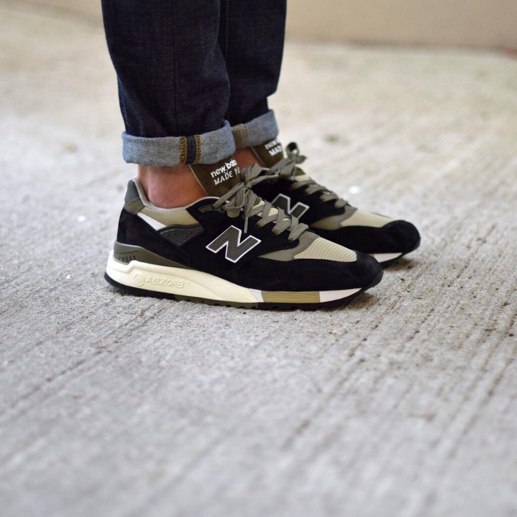 Black and tan New Balance athletic sneakers worn with dark denim jeans on concrete.