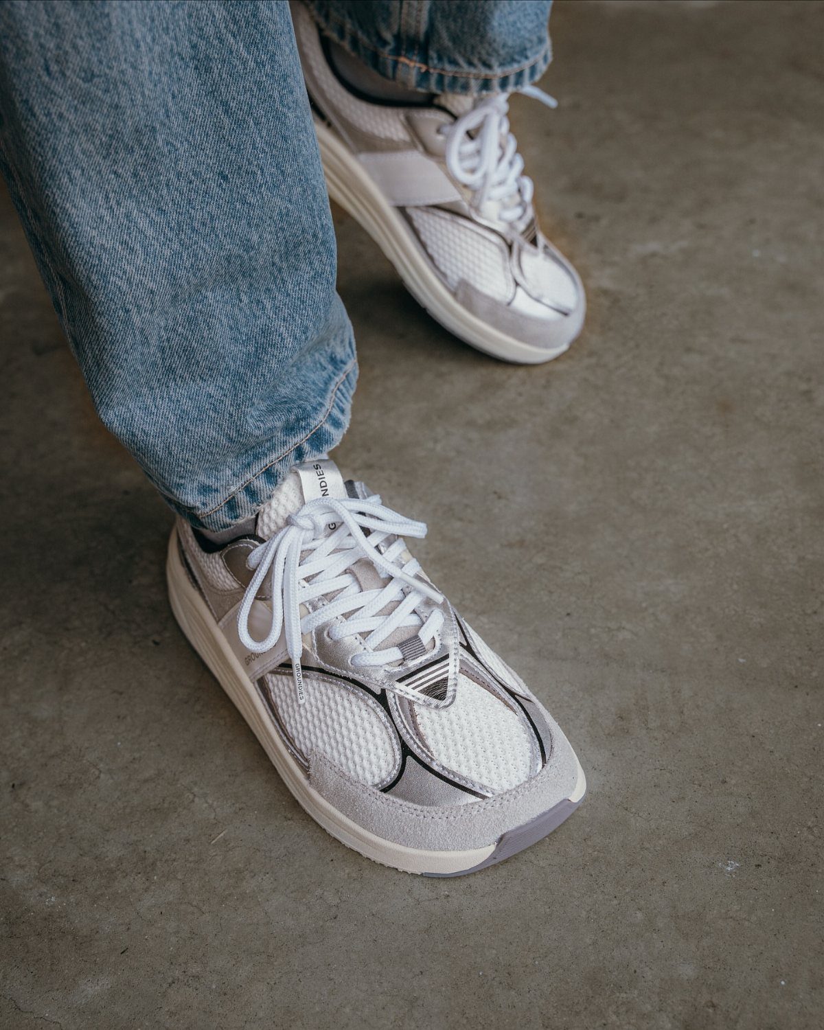 Light grey and white mesh sneakers worn with blue denim jeans on concrete.